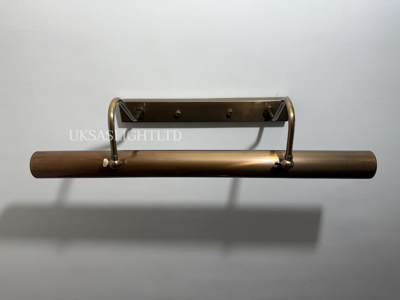 Op de afbeelding: Een bronzen schilderijlamp. De lamp heeft een cilindrisch lichaam met gebogen armen en een rechthoekige montageplaat. De tekst "UKSASLIGHTLTD" is zichtbaar op de muur.