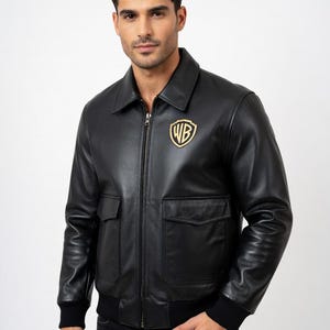 May include: Black leather jacket with a gold WB logo on the chest. The jacket has a collared neckline, two front pockets, and a ribbed hem and cuffs. The jacket is zipped up and has a classic bomber style.