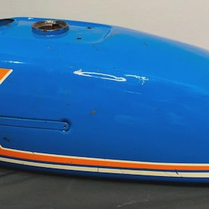Op de afbeelding: Een vintage blauwe motorbrandstoftank met oranje en witte strepen. De tank heeft een afgeronde vorm en vertoont tekenen van slijtage. Het ontwerp omvat een opening voor de tankdop.