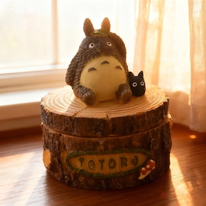 May include: A wooden box shaped like a tree stump, with a Totoro figurine and a small black cat on top. The box has the word "TOTORO" on the front. The Totoro figure is brown and cream-coloured, with a leaf on its head.
