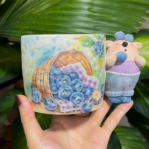 Puede incluir: Una taza de cerámica con un diseño de arándanos. La taza presenta una cesta rebosante de arándanos, una tela a cuadros morada y blanca y una linda figura de oso. La taza está pintada con detalles al estilo acuarela.
