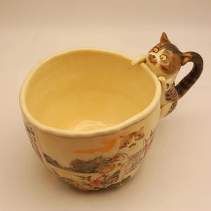 Puede incluir: Una taza de cerámica con un asa en forma de gato caprichoso. La taza es de color crema con un interior amarillo y presenta una ilustración detallada alrededor de la base. El asa del gato es marrón y blanca, lo que la hace única.