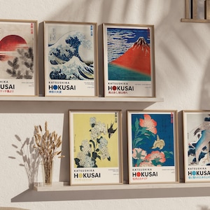 May include: A collection of six framed art prints by Katsushika Hokusai displayed on two shelves. The prints feature iconic Japanese woodblock designs including a red sun, a large wave, a red mountain, white flowers, orange flowers, and a crane.