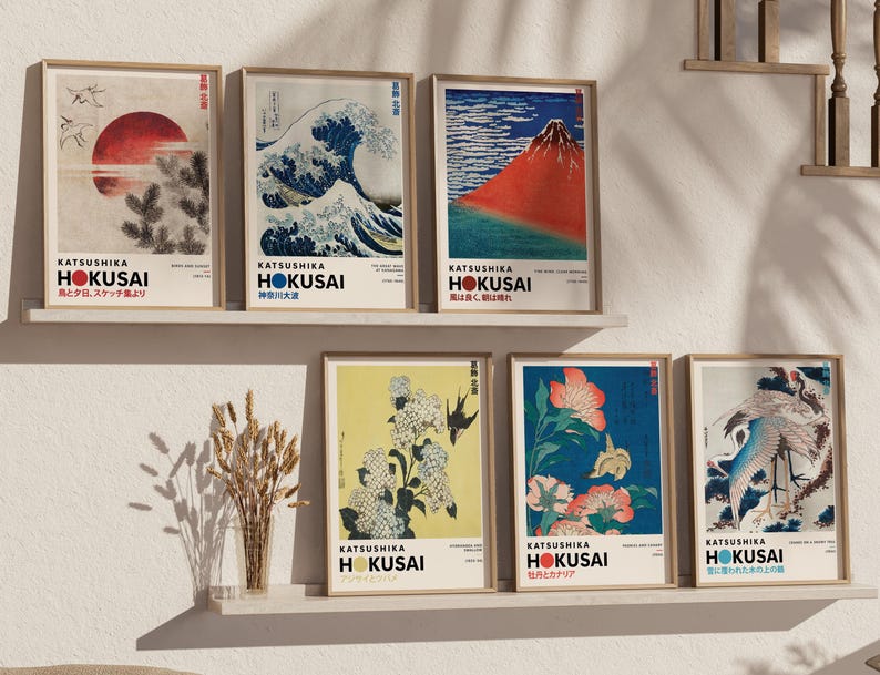 May include: A collection of six framed art prints by Katsushika Hokusai displayed on two shelves. The prints feature iconic Japanese woodblock designs including a red sun, a large wave, a red mountain, white flowers, orange flowers, and a crane.