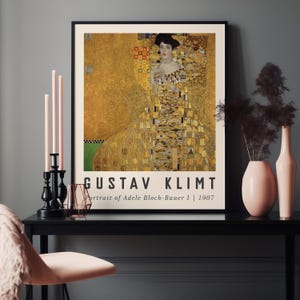 Puede incluir: Una impresión artística enmarcada del "Retrato de Adele Bloch-Bauer I" (1907) de Gustav Klimt. La obra presenta a una mujer con un vestido dorado y amarillo. La impresión se exhibe sobre una mesa negra con elementos decorativos.