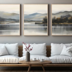 Könnte beinhalten: Drei gerahmte Landschaftskunst-Drucke, die eine ruhige See-Szene mit Bergen im Hintergrund zeigen. Das Kunstwerk verwendet gedämpfte Töne von Grau, Blau und Gold, wodurch eine ruhige Atmosphäre entsteht. Die Drucke werden über einem Holzsofa mit weißen Kissen ausgestellt.