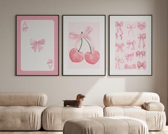May include: Three framed art prints with pink and white watercolor designs. The prints feature a playing card with a bow, cherries with a bow, and various pink bows. The frames are black, and the background is a neutral color.