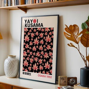 May include: A framed art print featuring the text "YAYOI KUSAMA" above a black background filled with pink maple leaf patterns. The print also includes Japanese text and "OSAKA 1992". The artwork is displayed on a white surface.