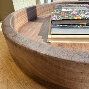 May include: A large, round wooden tray with a raised edge, filled with books. The tray is made of dark brown wood and sits on a light brown surface. The books have colorful floral covers and various titles.