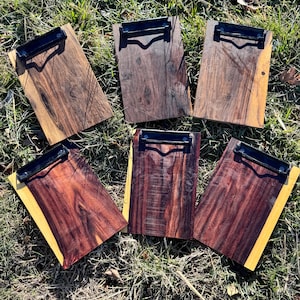 May include: Six wooden clipboards with black metal clips are arranged on a bed of grass. The clipboards have a rectangular shape with a natural wood grain finish. Some have a yellow border. The wood tones vary from light to dark brown.