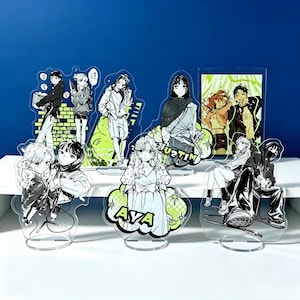 May include: A collection of seven clear acrylic character standees featuring anime-style illustrations in black, white, and lime green. The designs include various figures in casual clothing, some with text like AYA and MITSUKI, all mounted on circular bases.