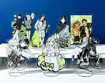 Può includere: Una collezione di sette espositori in acrilico trasparente con illustrazioni in stile anime in bianco, nero e verde lime. I design includono varie figure in abbigliamento casual, alcune con il testo AYA e MITSUKI, montate su basi circolari.