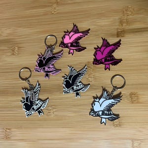 May include: Keyrings with a bird design and the text "Cry Baby." The keyrings are in black, white, and pink, with silver rings. The bird has outstretched wings, and the text is on a banner below.