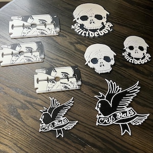 May include: A selection of decorative items with black and white designs. Includes razor blade-shaped pieces with a face illustration, skull-shaped pieces with the text "Suicideboys", and bird-shaped pieces with the text "Cry Baby".