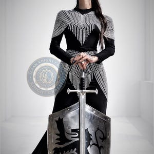 Può includere: Una persona in un abito nero e un'armatura a maglia di catena tiene una spada e uno scudo. Lo scudo ha un emblema di leone nero. L'armatura comprende un mantello e una cintura a maglia di catena. Tema medievale.