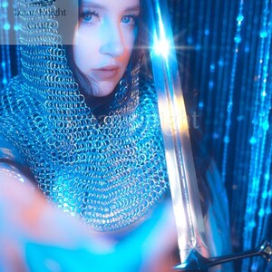 May include: A person wearing a chainmail hood and holding a sword. The sword is silver with a detailed hilt. The image has a blue and white colour scheme, with the text "Izaar Knight Crafts" visible in the upper left corner.