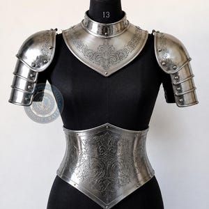 May include: A polished steel breastplate and gorget set featuring intricate floral engravings. The armour includes matching pauldrons with layered plates and rivets, displayed on a black mannequin torso. The metal surface reflects light.