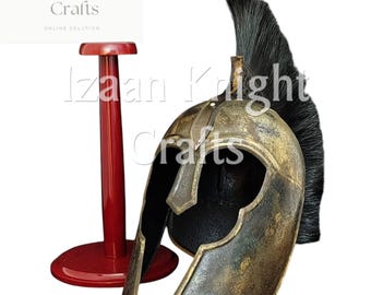 May include: A bronze-colored helmet with a black plume, displayed next to a red stand. The helmet has a weathered appearance and is likely a replica. The logo for Izaan Knight Crafts is in the upper left corner.
