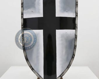 May include: A medieval shield with a black cross on a silver background. The shield has a metal frame with rivets. The shield is displayed on a wooden stand. The shield is a replica of a historical design.