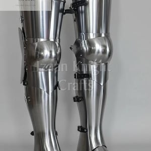 May include: A pair of polished steel leg armor, featuring articulated plates and straps. The armor is designed to protect the legs and feet. The image includes the text "Izaan Knight Crafts" and "ONLINE SOLUTION".