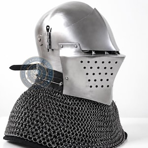 Op de afbeelding: Een gepolijste stalen bekkenhelm met een scharnierend vizier voorzien van een reeks ventilatiegaten. De helm is bevestigd aan een donkere maliënkolder die over de schouders valt. Een cirkelvormig logo met de tekst Knight Crafts is zichtbaar.