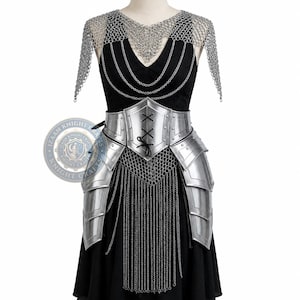 May include: A black dress adorned with silver-toned metal armour, featuring a chainmail shoulder mantle, a front-lacing corset-style waist piece, layered hip plates, and cascading chain fringe. The armour includes a circular logo reading IZAAN KNIGHT CRAFTS.