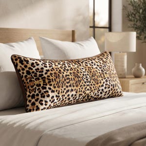 May include: A rectangular lumbar pillow featuring a prominent leopard print pattern in shades of brown, black, and tan. The pillow has a dark brown piped edge and is positioned horizontally in front of two solid white pillows on a bed.