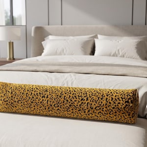 May include: A cylindrical bolster pillow featuring a vibrant yellow and black leopard print pattern. The pillow is positioned horizontally on a white upholstered bench at the foot of a bed with white bedding and a neutral-toned headboard.