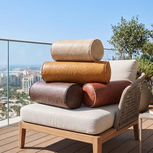 Puede incluir: Una pila de cuatro cojines cilíndricos en varios tonos de beige, marrón claro, marrón oscuro y marrón rojizo, apoyados sobre un sillón de exterior de color claro con respaldo trenzado y estructura de madera en una terraza.