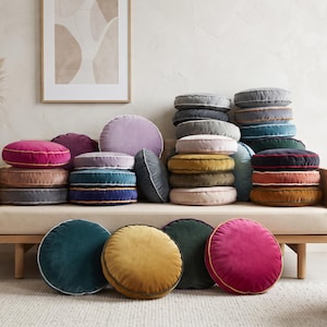 May include: A collection of round velvet cushions in various colours, including pink, mustard, teal, grey, and purple. Each cushion features a piped edge detail. The cushions are stacked on a wooden daybed and placed on the floor in front of it.
