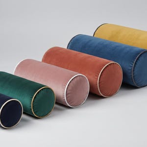 May include: A set of five cylindrical bolster pillows in varying sizes and colors, including navy, emerald green, dusty pink, terracotta, and mustard yellow. Each velvet pillow features contrasting piping along the circular end edges.