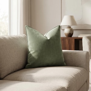May include: A square throw pillow in a solid sage green color with a smooth, velvet-like texture and piped edges. The pillow is positioned upright on a light beige sofa, featuring a distinct pinched top corner that creates a structured shape.