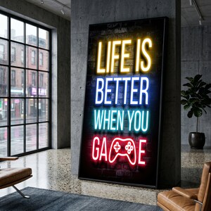 Op de afbeelding: Een verticaal rechthoekig wandkunstwerk met neon-stijl tekst op een donkere bakstenen achtergrond. De tekst luidt "LIFE IS" in geel, "BETTER" in blauw, "WHEN YOU" in lichtblauw en "GAME" in rood met een wit gamecontroller-icoon.