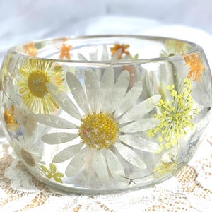 May include: A clear, round glass bowl with embedded dried flowers. The bowl features large white daisies with yellow centres, along with smaller yellow and orange flowers. The flowers are suspended in the clear glass, creating a decorative effect.