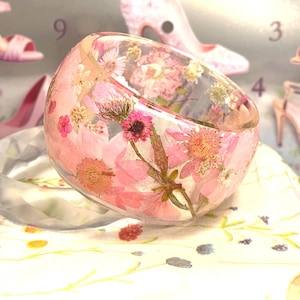 Könnte beinhalten: Ein Armreif aus klarem Harz mit eingebetteten getrockneten rosa und weißen Blumen. Das kreisförmige Accessoire zeigt eine Vielzahl von Blumenformen und grünen Stielen, die im transparenten Material schweben und ein botanisches Muster auf dem gesamten Band erzeugen.