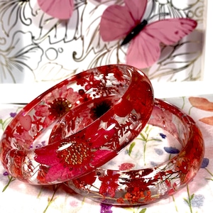 May include: Two clear resin bangle bracelets featuring embedded dried red flowers and small white beads. The circular bracelets have a smooth, glossy finish, showcasing a variety of vibrant red floral petals and greenery suspended within the transparent material.