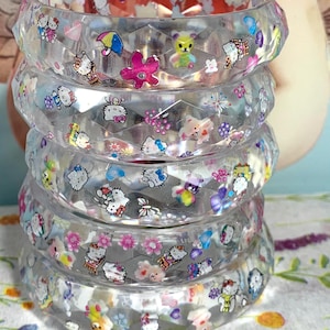 Op de afbeelding: Een stapel van vijf doorzichtige, gefacetteerde harsarmbanden met ingebedde kleurrijke stickers van Hello Kitty-figuren, kleine teddyberen, bloemen, hartjes, paraplu's en vlinders. De armbanden hebben een geometrische textuur met meerdere zijden.