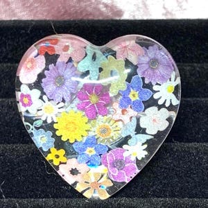 May include: A heart-shaped resin pendant filled with colourful floral designs. The pendant features a variety of flowers in shades of pink, purple, yellow, and blue, set against a clear background. A unique piece of jewellery.