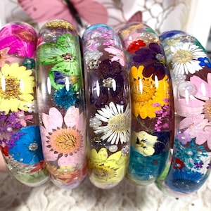 May include: Five clear resin bangles featuring embedded dried flowers in various colours including yellow, pink, blue, purple, and green. Each bracelet contains a unique arrangement of pressed petals, small white beads, and glitter accents.
