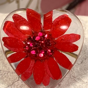 May include: A heart-shaped clear resin ring featuring a vibrant red flower with elongated petals embedded inside. The center of the flower is filled with small, reflective red glitter particles that catch the light, creating a bright, textured appearance.
