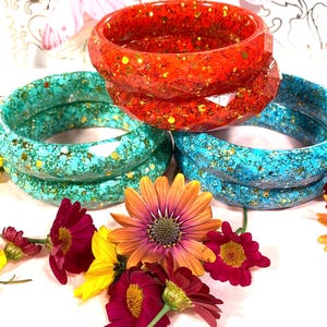 May include: A collection of six faceted resin bangles in vibrant red, turquoise, and blue, each embedded with shimmering gold glitter. The bangles are arranged in a stack and surrounded by colourful fresh flowers including orange, yellow, and deep pink.
