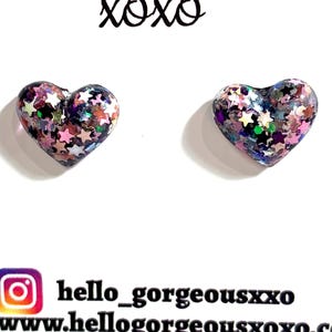 May include: A pair of heart-shaped earrings with a clear base, filled with colourful glitter and star-shaped confetti. The text "Hello Gorgeous xoxo" is above the earrings. The earrings are on a white background.