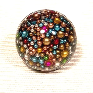 Könnte beinhalten: Ein kreisförmiger Cabochon mit einer dichten Ansammlung kleiner, mehrfarbiger, kugelförmiger Perlen in Gold, Metallic-Blau, leuchtendem Pink, Silber und Grün, die alle in einer klaren, glatten und abgerundeten Oberfläche aus Glas oder Harz eingeschlossen sind.