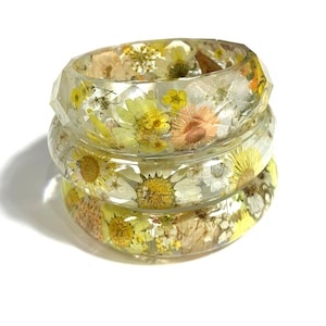 May include: Three clear resin bangles with embedded dried flowers in shades of yellow, white, and peach. The bangles are stacked, showcasing the floral details. The faceted design catches the light, highlighting the natural elements.