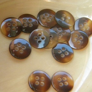 May include: A collection of brown and black buttons with four holes each. The buttons are arranged in a semi-circle on a light brown surface.