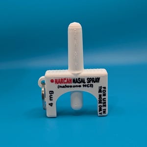 May include: A white 3D-printed plastic keychain replica of a Narcan nasal spray device. The text reads NARCAN NASAL SPRAY (naloxone HCl), 4 mg, and FOR USE IN THE NOSE ONLY. A silver metal keyring is attached to the side of the white plastic housing.