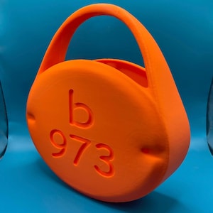 May include: Bright orange, rounded handbag with a curved handle. The bag features the letter "b" and the number "973" embossed on the front. The bag is made of a solid material and has a smooth texture.