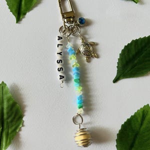 Puede incluir: Un llavero hecho a mano con el nombre "ALYSSA" escrito con cuentas de letras blancas. El llavero presenta un cierre dorado, un pequeño dije de tortuga y un grupo de cuentas azules, verdes y amarillas. Una esfera amarilla está encerrada en una jaula de alambre plateado.