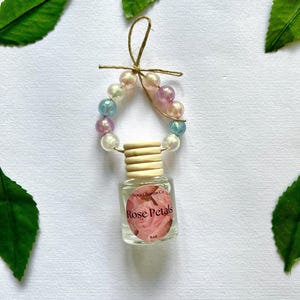 Puede incluir: Un pequeño frasco de vidrio para ambientador de coche con tapón de madera, suspendido por un lazo de cuerda adornado con cuentas pastel iridiscentes en tonos rosa, azul y blanco. La etiqueta muestra una imagen de rosa y el texto Nova Charms Co Rose Petals 8ml.
