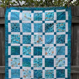 May include: A handmade quilt with a sea life theme. The quilt features a grid pattern with squares of various blue fabrics, including wave patterns, fish, and jellyfish. White borders and a teal border frame the quilt.